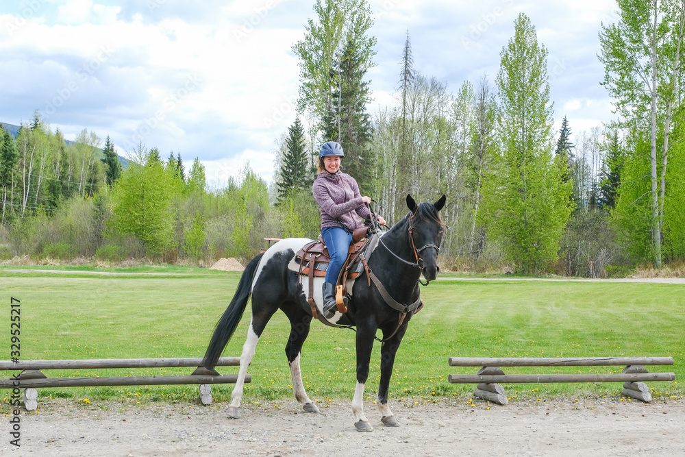 A young woman riding a black and white horse in front of a green field, Banff national park