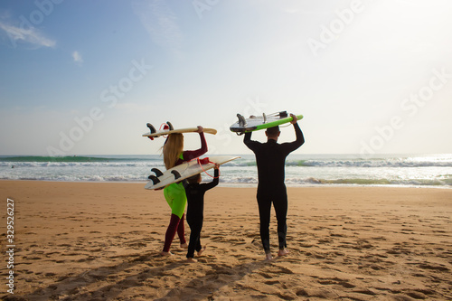 Family with surfboards walking on beach. Back view of parents with cute little son in wetsuits holding surfboards and walking to ocean waves. Surfing concept