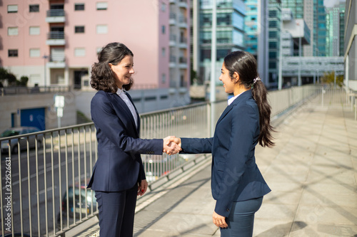 Content businesswomen shaking hands. Side view of cheerful female colleagues in formal wear standing on street and greeting each other. Partnership concept