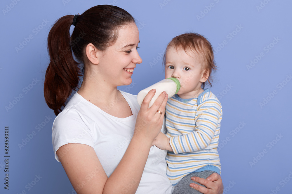 Close up photo of positive good looking young brunette, having ponytail, feeding child with milk from bottle, smiling sincerely, standing isolated over blue background in studio. Childhood concept. Close up photo of positive good looking young brunette, having ponytail, feeding child with milk from bottle, smiling sincerely, standing isolated over blue background in studio. Childhood concept.