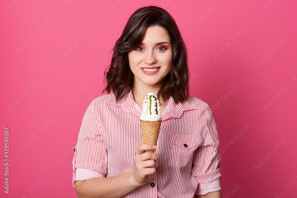 Image of pleasant sincere good looking brunette holding big portion of ice cream with jam, standing isolated over pink background, wearing striped shirt, having short black hair. Tasty food concept.