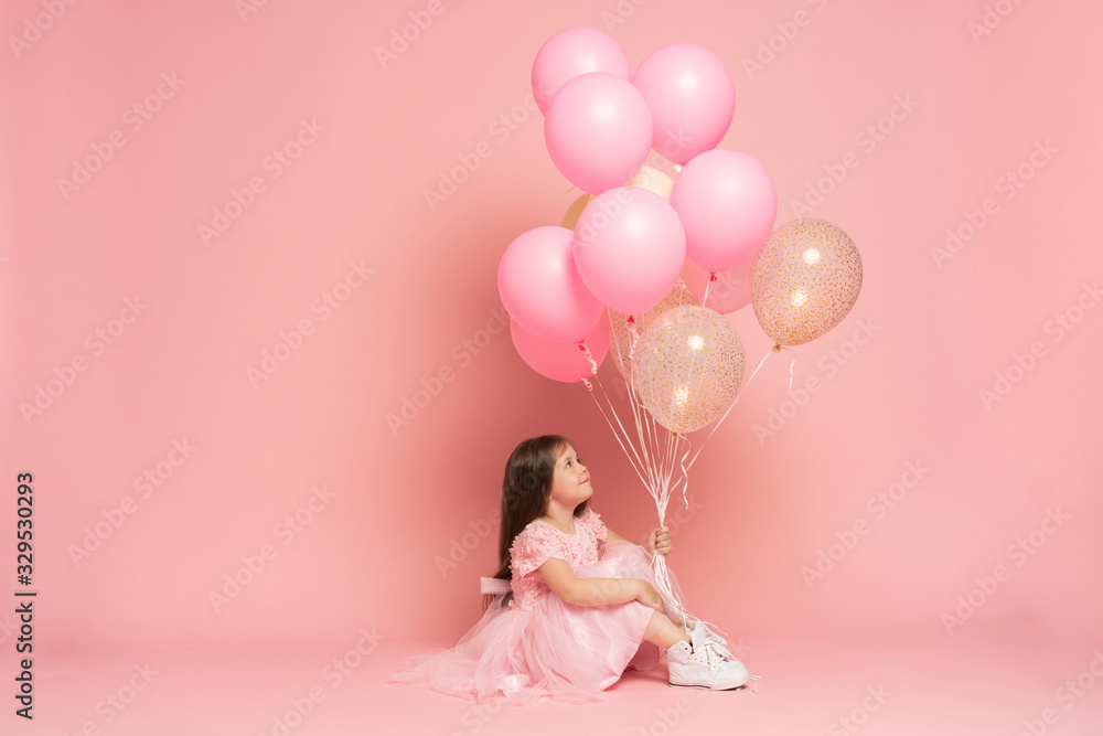 Happy celebration of birthday party with flying balloons of charming cute little girl in tulle dress smiling to camera isolated on pink background. Charming smile, expressing happiness