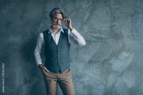 Portrait of pensive minded elegant rich wealthy old macho man touch his modern spectacles put hands pocket wear good look brown pants trousers isolated over grey color background © deagreez