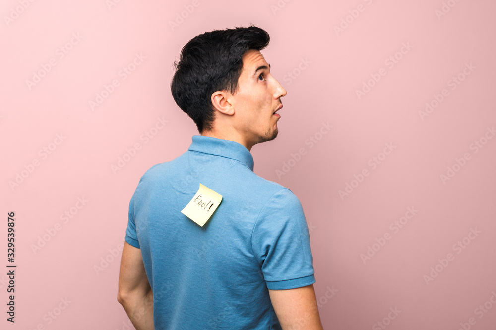 fool's day. foolish looking man in blue polo shirt with a yellow sticky note with word fool on his back stupidly looking up against pink background