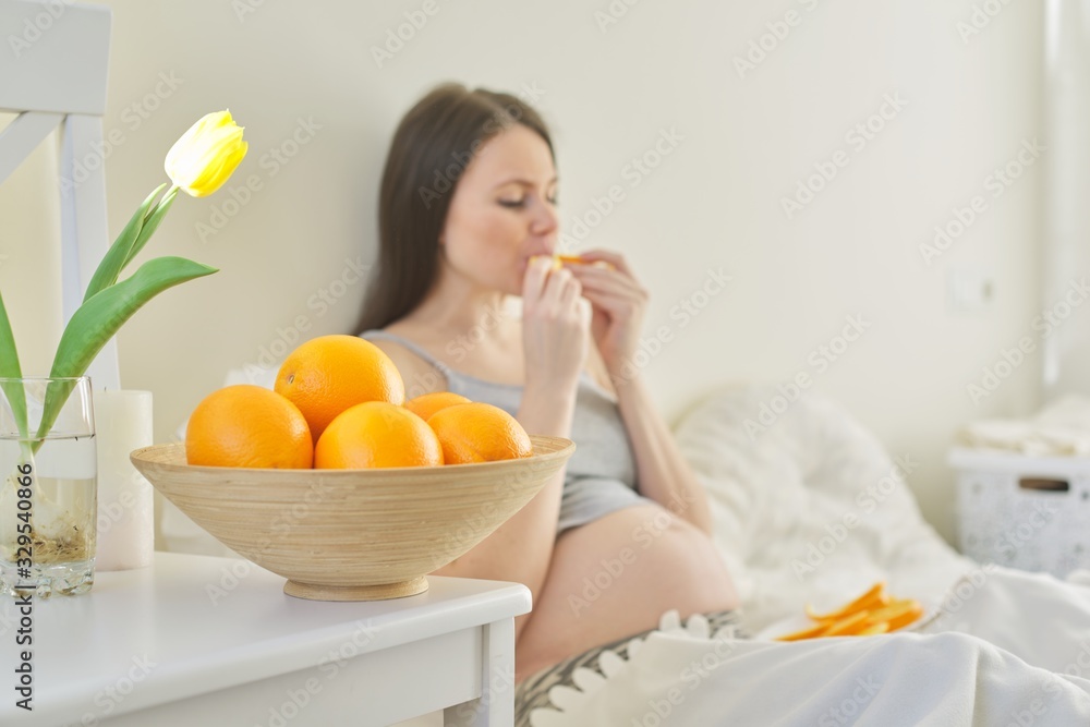 Bowl with oranges in focus, pregnant young woman eating slices of oranges sitting in bed Bowl with oranges in focus, pregnant young woman eating slices of oranges sitting in bed