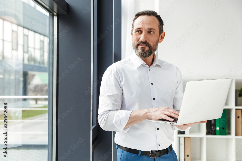Photo of bearded pleased businessman in white shirt typing on laptop