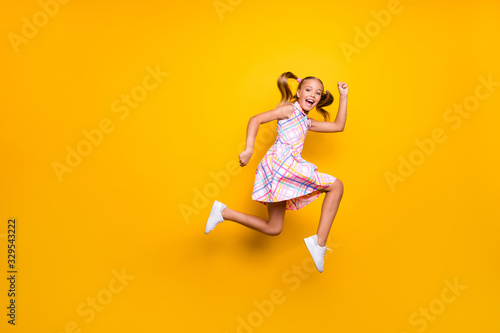 Full size profile side photo of cheerful crazy funny small kid girl jump run after spring time discount wear checkered skirt footwear isolated bright yellow color background