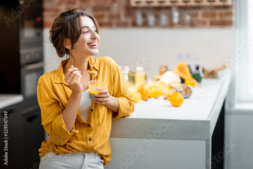 Portrait of a young and cheerful woman eating chia pudding, having a snack or breakfast in the kitchen with lots of fresh fruits and vegetables. Concept of vegetarianism, healthy eating and wellness