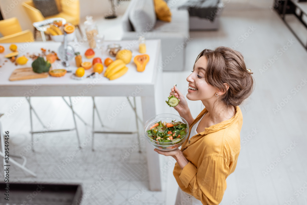 Portrait of a young and cheerful woman dressed in bright shirt eating salad at home. Concept of wellbeing, healthy food and homeliness