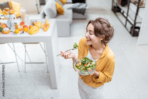 Portrait of a young and cheerful woman dressed in bright shirt eating salad at home. Concept of wellbeing, healthy food and homeliness