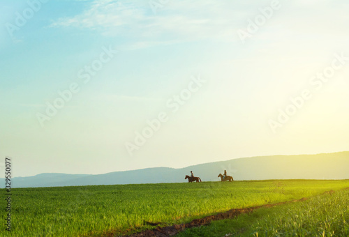 two distant horse riders having a ride under spring bright blue sky in green countryside field against mountains silhouette