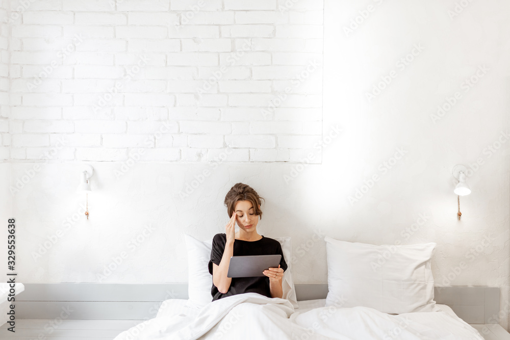 Young woman with a digital tablet lying in bed, wide interior view of a white bedroom. Concept of a leisure time with mobile wireless devices at home