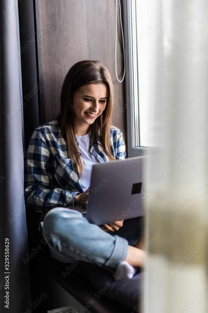 Young pretty girl sitting near the window with a laptop at home on the windowsill Young pretty girl sitting near the window with a laptop at home on the windowsill