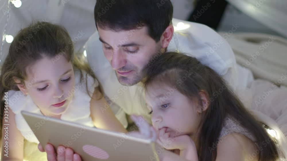 Father in White Shirt and Two Little Girls in a White Dresses Lies on the Carpet and Plays with a Tablet Computer in Christmas Decoration. New Year Family Holiday. The Concept of a Parenthood, Family. Father in White Shirt and Two Little Girls in a White Dresses Lies on the Carpet and Plays with a Tablet Computer in Christmas Decoration. New Year Family Holiday. The Concept of a Parenthood, Family.