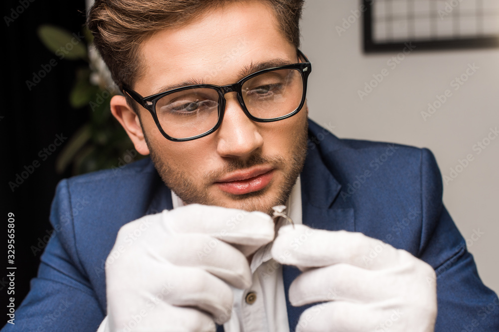 Selective focus of handsome jewelry appraiser in gloves holding ring with gemstone in workshop Selective focus of handsome jewelry appraiser in gloves holding ring with gemstone in workshop
