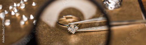 Selective focus of magnifying glass, jewelry ring with gemstone in tweezers on board, panoramic shot photo