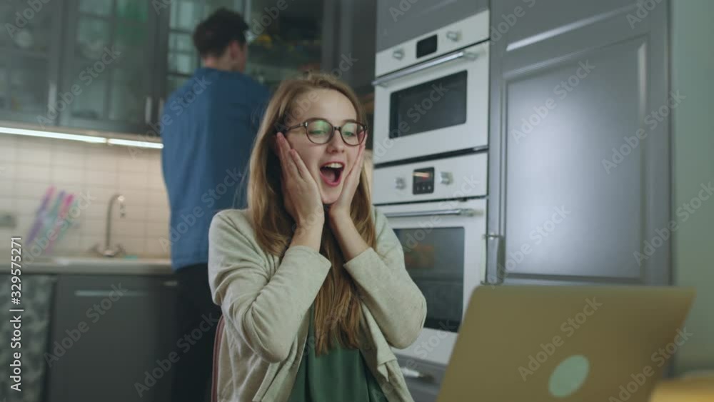 Young Attractive Girl Using Laptop While Sitting in Kitchen with Guy in Background. She Learns Good News Making Her Very Happy and He Approaches Her to Congratulate and Hug. Slow Motion Shot