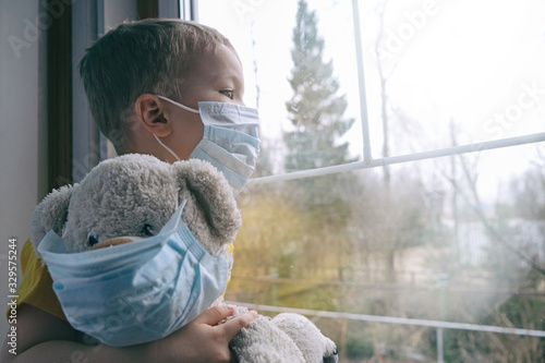 Sad illness child on home quarantine. Boy and his teddy bear both in protective medical masks sits on windowsill and looks out window. Virus protection, coronavirus pandemic, prevention epidemic.