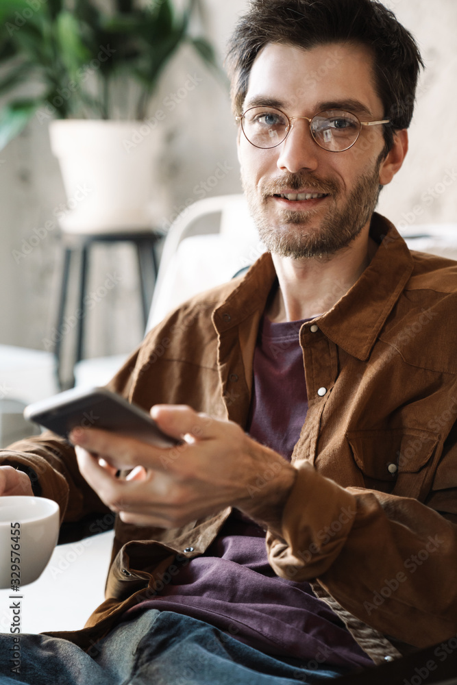 Image of young man in eyeglasses drinking tea and using cellphone in cafe Image of young man in eyeglasses drinking tea and using cellphone in cafe