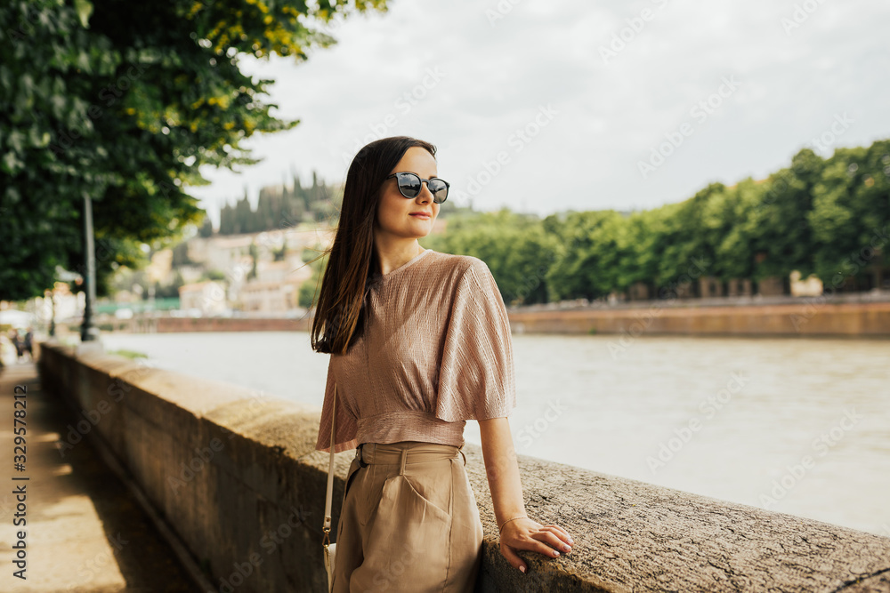 Portrait of a brunette stylish  woman sitting on the stone fence by the river in city of Verona, Italy. Concept of travel, destination, summer, vacation.