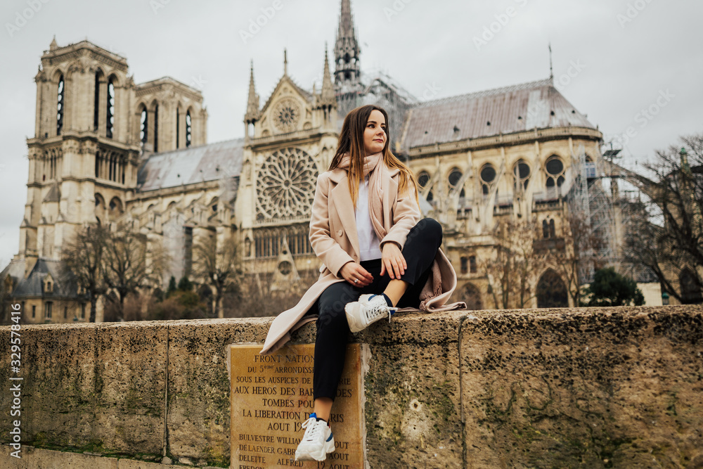 Young stylish woman tourist sitting near the famous Notre Dame cathedral in Paris, France. Handsome girl sitting in front of the Notre Dame cathedral in Paris at the bridge.