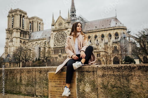 Obraz na płótnie Young stylish woman tourist sitting near the famous Notre Dame cathedral in Paris, France. Handsome girl sitting in front of the Notre Dame cathedral in Paris at the bridge. w salonie