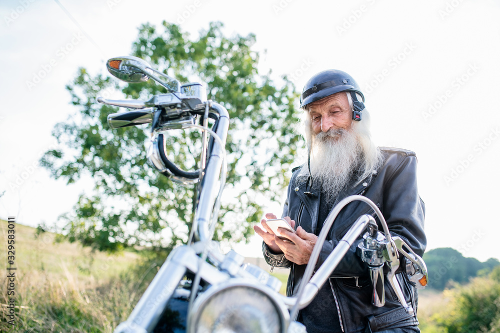 Senior man traveller with motorbike in countryside, using smartphone.