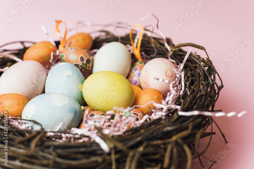 A nest of twigs and hay and a pink paper filler with pastel colored Easter eggs. Painted and decorative eggs for a light Easter holiday. Easter card on a pink background