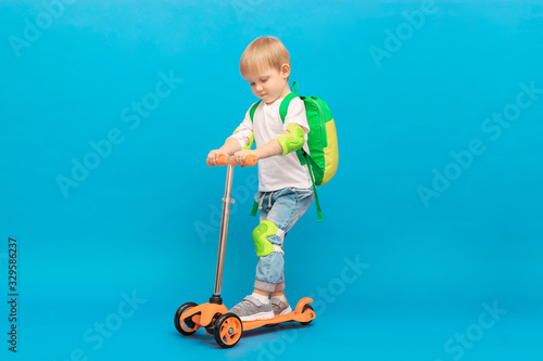 A blond-haired boy of 4 years old, a child with a backpack and in jeans with a white T-shirt rides on an orange scooter with light green protection on his elbows and knees.