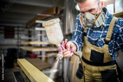 Carpenter with paint spray gun painting wooden plank in carpentry workshop