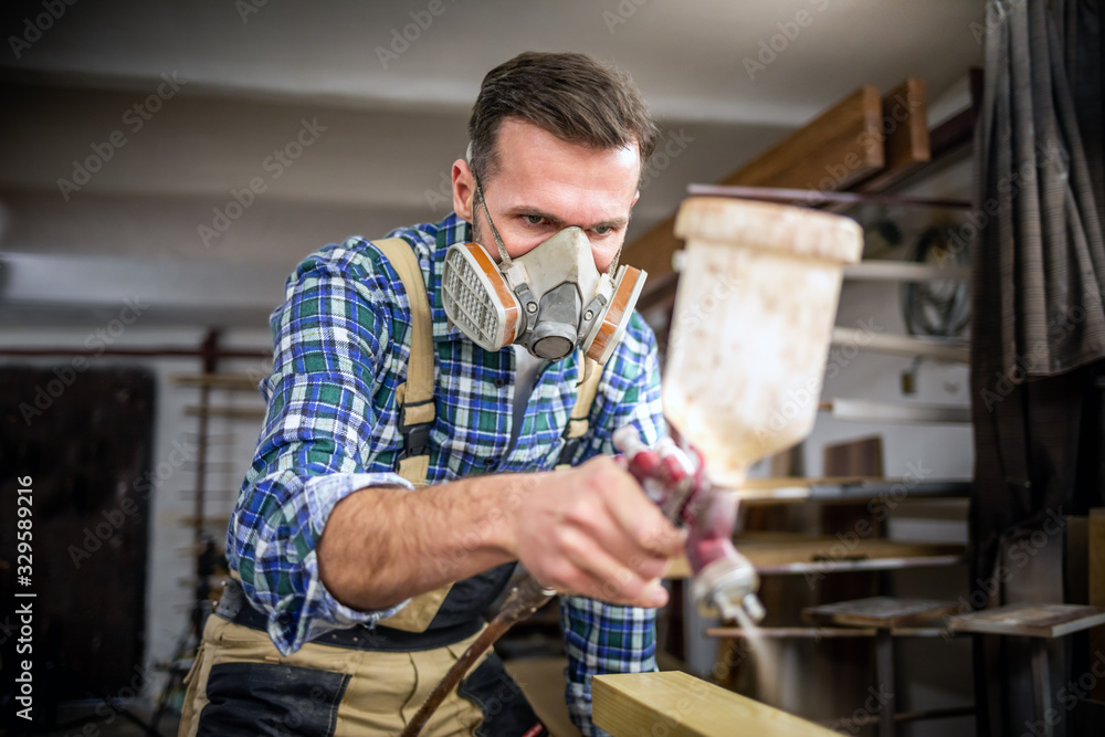 Carpenter with mask using paint spray gun to painting wooden plank in carpentry workshop