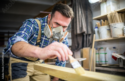 Carpenter with mask applies paint using paintbrush in carpentry workshop