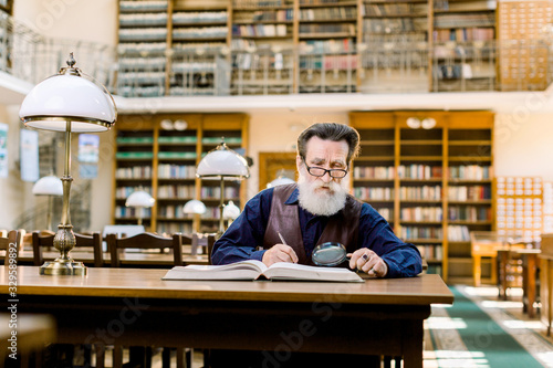 Senior man working in old library, sitting at the table with books and magnifying glass, making notes. Student old man, education in library, old shelves with books on the background