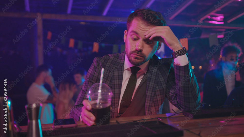 Handsome tired businessman sitting at bar counter drinking alcohol thinking over life with upset expression at night youth party in the club.