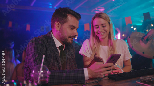 Handsome businessman and a happy girl laughing of funny memes on smartphone sharing exciting stories chilling together at bar counter.
