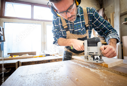 Carpenter holds milling machine and and working at wooden board in woodworkin...