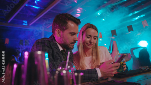 Attractive young couple looking through funny photographs on smartphone sitting at bar counter enjoying weekend party time together.