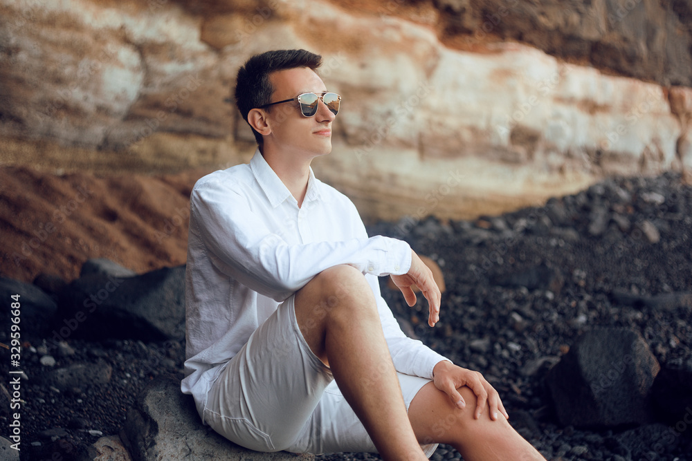 A man sits on the ocean on the rocks. Stone Beach in Tenerife. A man dressed in white, wearing glasses, looks into the distance. A man is relaxing on the beach, pondering, calm, muse. A man sits on the ocean on the rocks. Stone Beach in Tenerife. A man dressed in white, wearing glasses, looks into the distance. A man is relaxing on the beach, pondering, calm, muse.