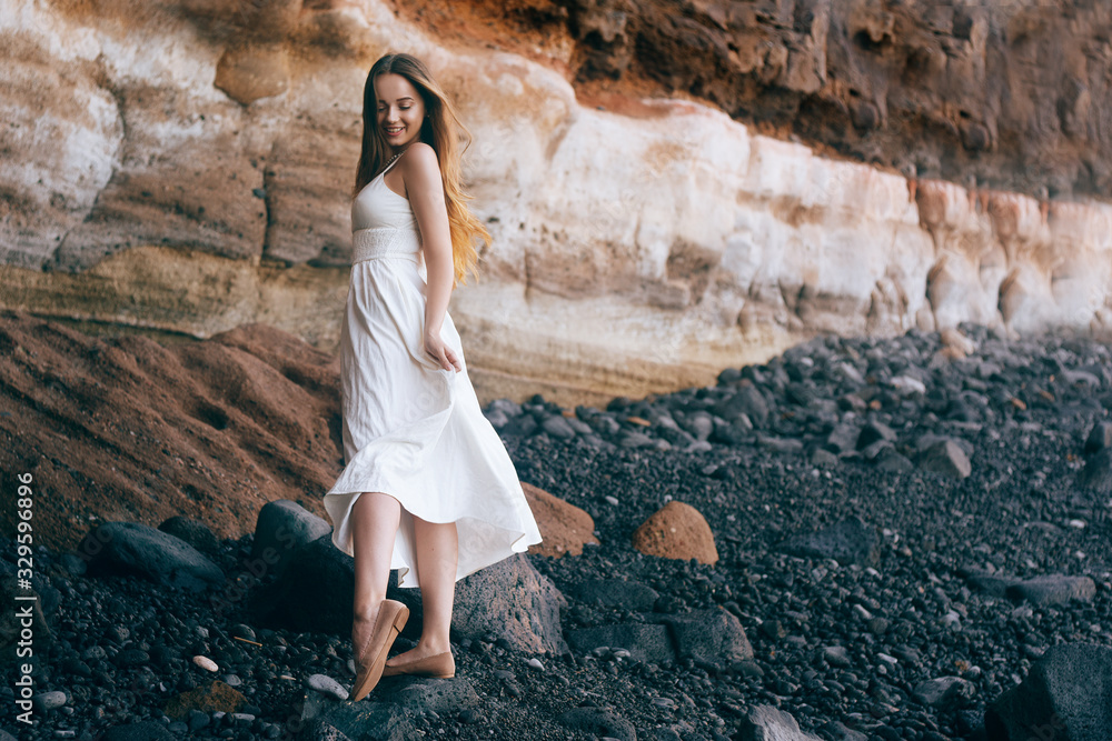 Gorgeous girl in a long white dress on the ocean at the beach. The long-haired girl is inherited by nature, mountains, stones, the beach. Girl on vacation, light dress, summer, date. Bride Gorgeous girl in a long white dress on the ocean at the beach. The long-haired girl is inherited by nature, mountains, stones, the beach. Girl on vacation, light dress, summer, date. Bride