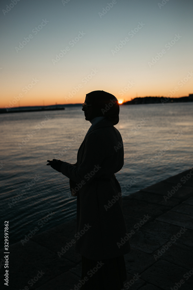 Fototapeta premium Silhouette of young European Muslim woman with hijab walking by the sea during sunset/sunrise time. Winter time.