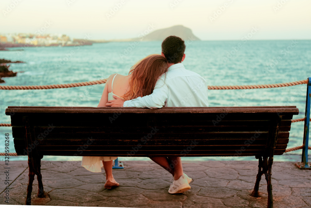 The concept of rest, vacation, travel, honeymoon. A girl and a guy are sitting on a bench, laughing against the backdrop of the ocean and mountains. Spain, Canaries.