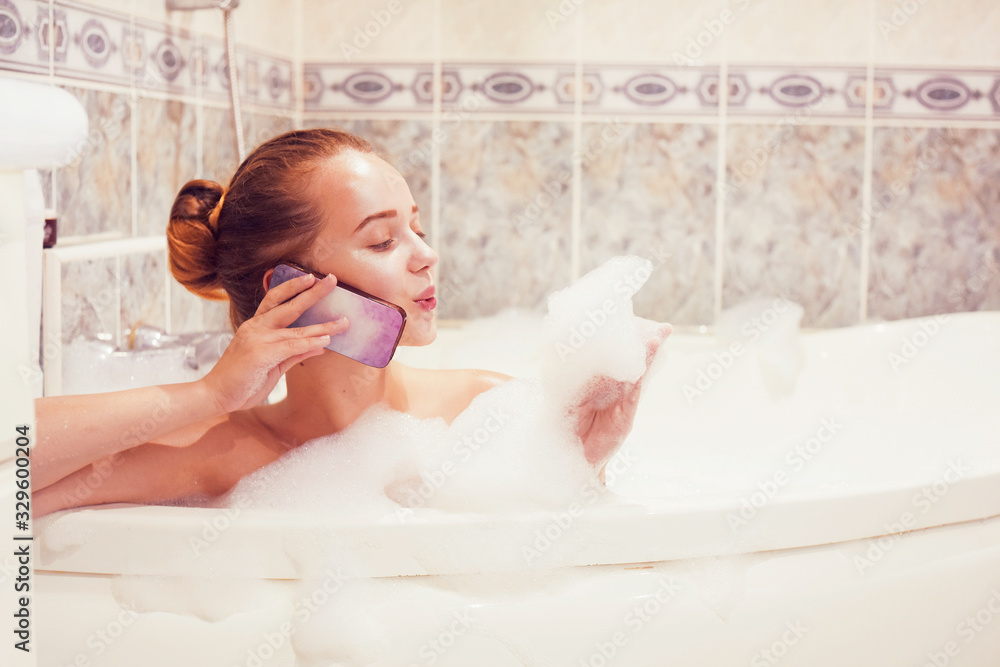 Naklejka premium The girl is lying in the whirlpool with foam and talking on a waterproof phone. The girl relaxes and gossips. Poetz girl in the foam. Conversation with a girlfriend.