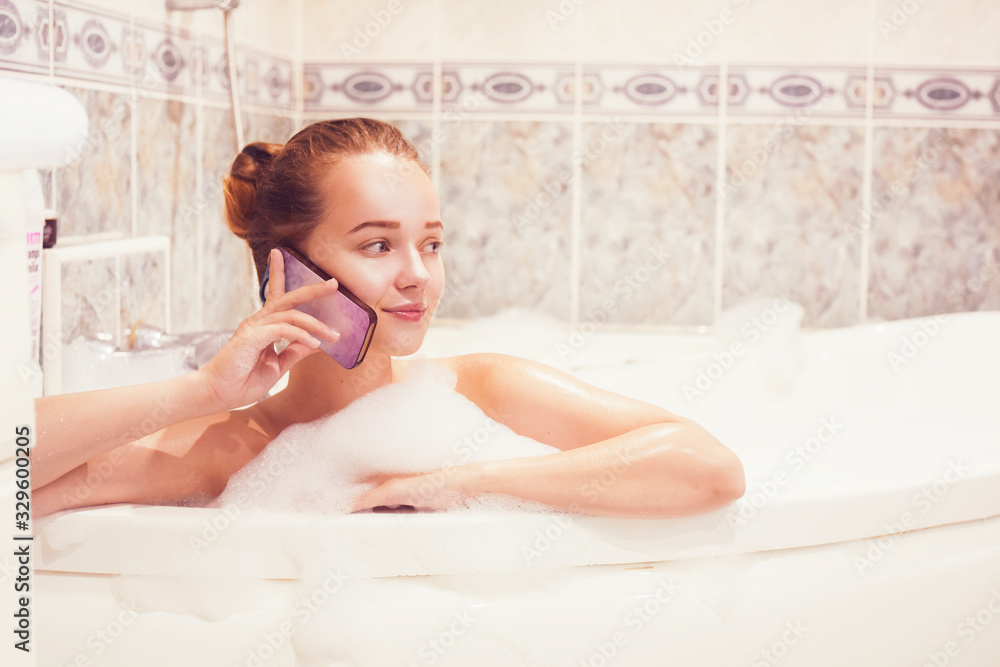 Obraz premium The girl is lying in the whirlpool with foam and talking on a waterproof phone. The girl relaxes and gossips. Poetz girl in the foam. Conversation with a girlfriend.
