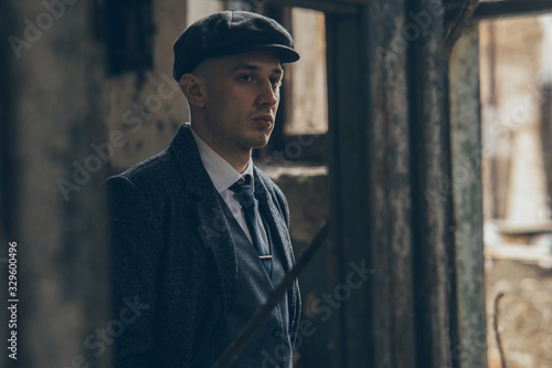 A man posing in the image of an English retro gangster dressed in a coat, suit and flat cap.