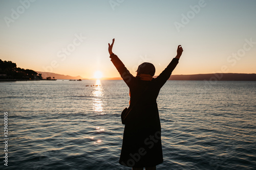 photo of young European Muslim women with hijab standing and looking into sea. She is standing turned back with her hands in the air. She is happy and relaxed. Photos taken during sunrise.   © Melika