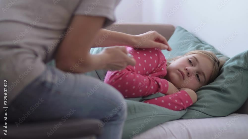 Mother is giving digital thermometer to her cold little daughter, lying on a couch in home Mother is giving digital thermometer to her cold little daughter, lying on a couch in home
