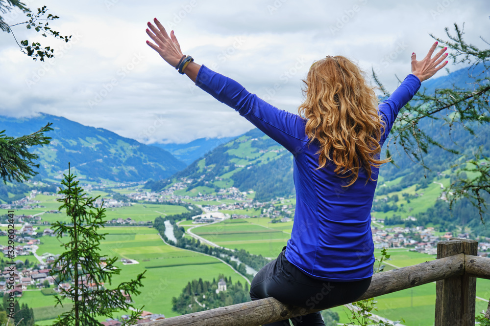Happy woman with arms spread out in joy and curly hair looks towards Zillertal valley in Austria, from a viewpoint high above the green valley.