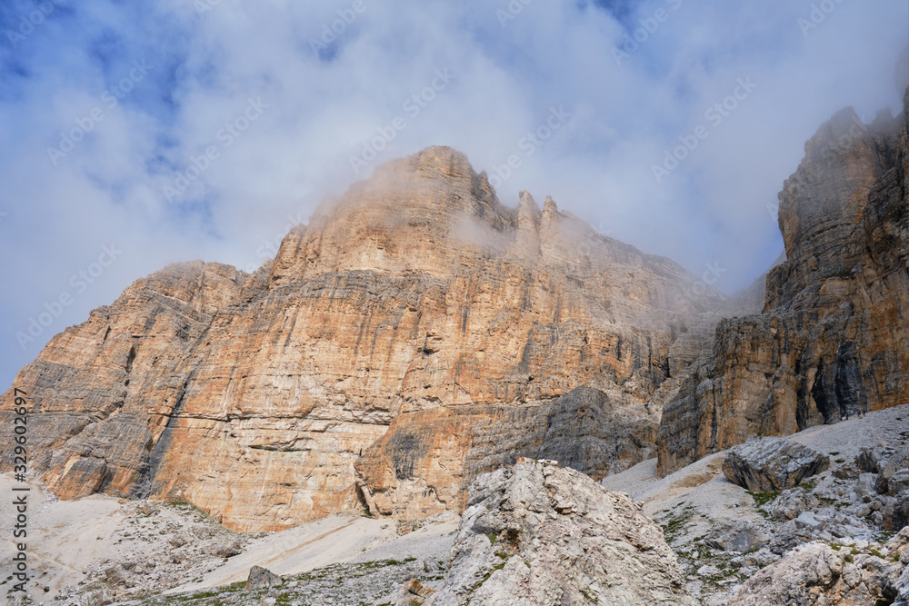 Approach to Cesare Piazzetta via ferrata start, with impressive Dolomites rock walls lit by sunlight, rock scree at the bottom, and clouds passing by.