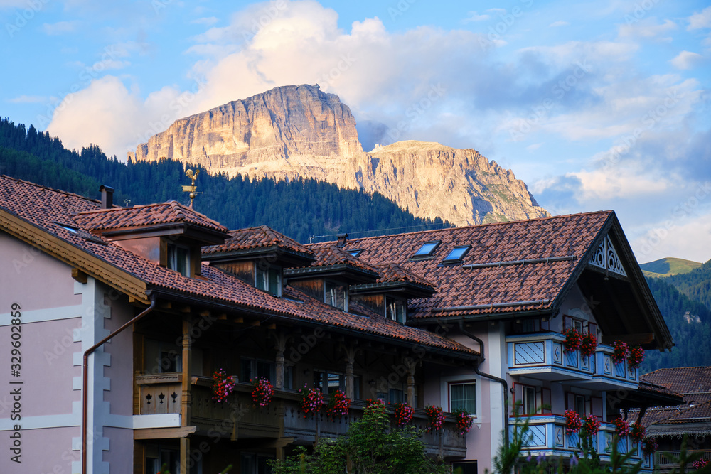 Traditional Italian house in Campitello di Fassa, with Dolomites mountain peaks lit by orange sunset light in the background.