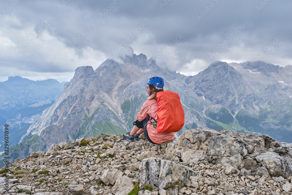 Woman on Colac peak, at the end of via ferrata dei Finanzieri, with helmet on and a backpack with rain cover, enjoys the panoramic views, with storm clouds approaching.